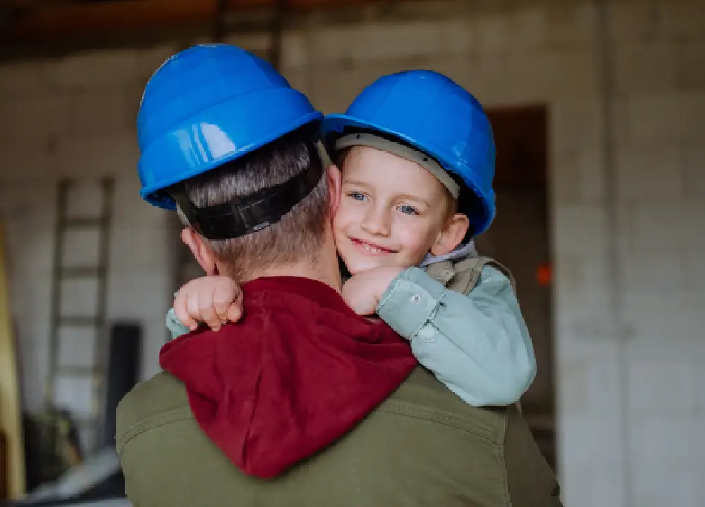 A young boy in a blue hard hat hugging his father on a construction site, illustrating the risk of secondary asbestos or talc exposure.