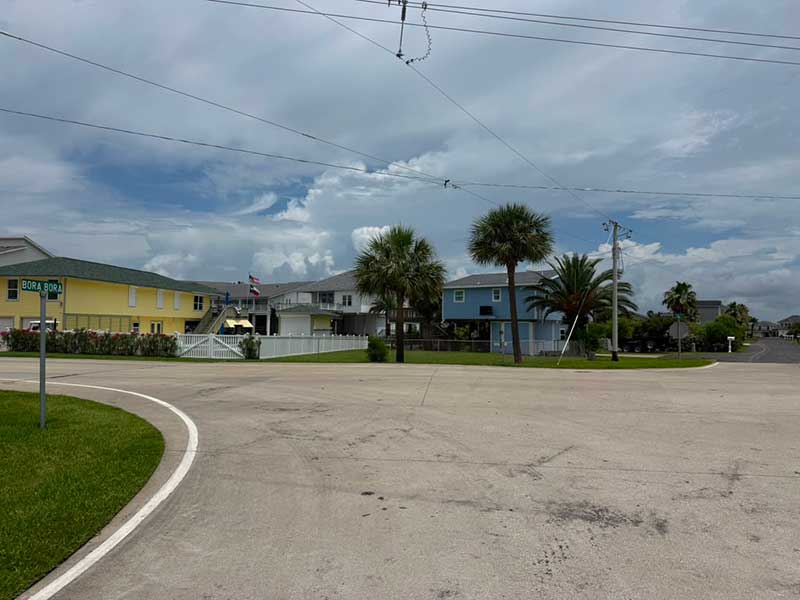Residential street corner on Tiki Island in Galveston, Texas, with elevated coastal homes, palm trees, and a Bora Bora street sign