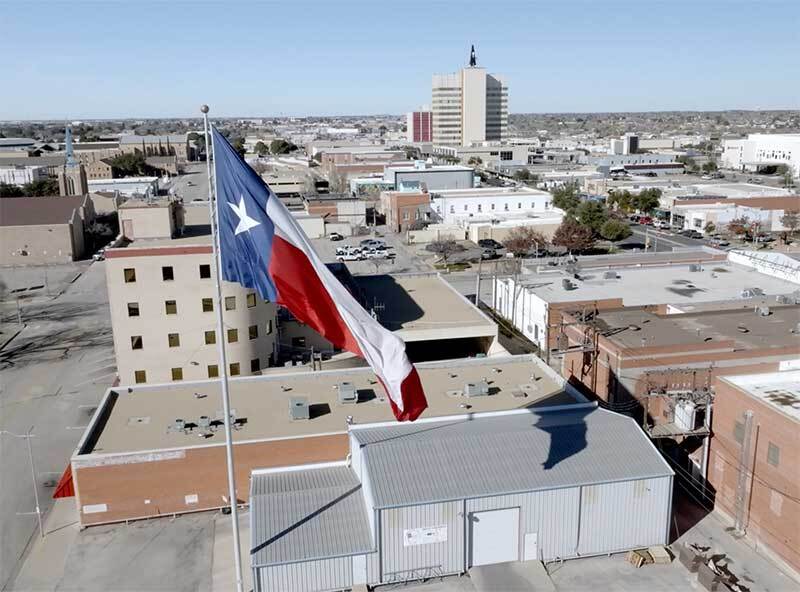 A large Texas flag flying over downtown rooftops, with an office tower in the distance under a clear blue sky